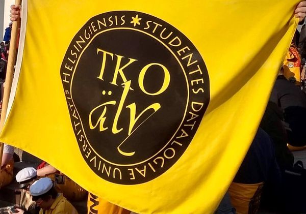TKO-äly's flag nearly filling the frame, some glimpses of overalls and people visible in the background