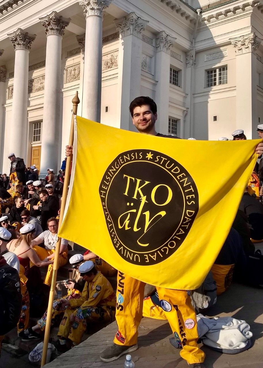 Sean Weber holding TKO-äly's flag and wearing overalls, Helsinki Cathedral and many students in overalls in the background